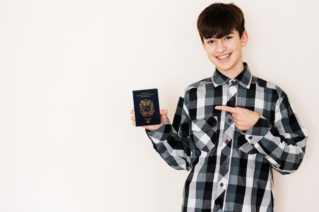 Garoto adolescente segurando o passaporte da Venezuela parecendo positivo e feliz de pé e sorrindo com um sorriso confiante contra o fundo branco