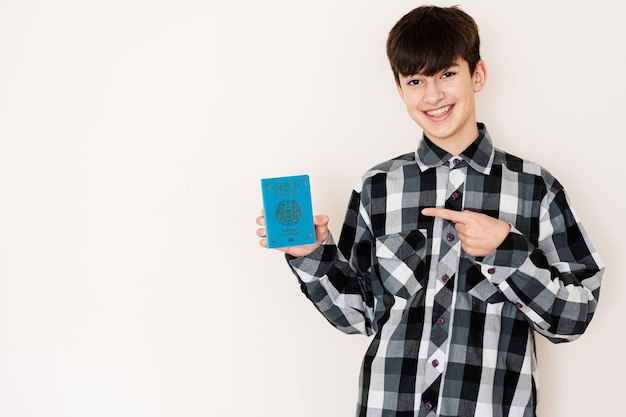 Garoto adolescente segurando o passaporte do Cazaquistão parecendo positivo e feliz em pé e sorrindo com um sorriso confiante contra o fundo branco