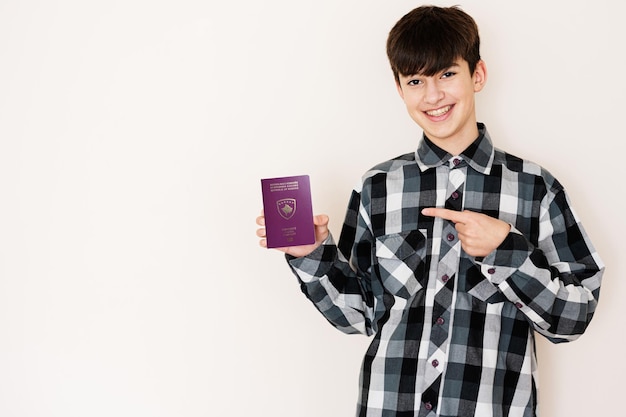 Garoto adolescente segurando o passaporte do Kosovo parecendo positivo e feliz em pé e sorrindo com um sorriso confiante contra o fundo branco