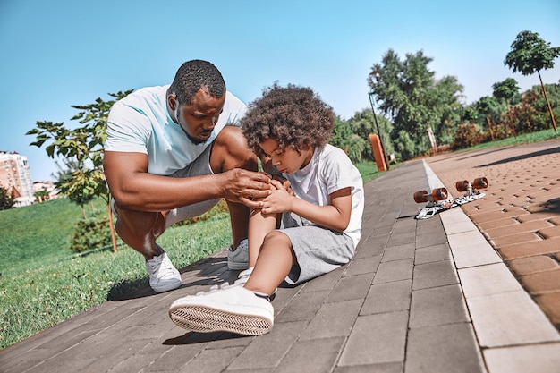 Garoto afro-americano e seu pai parecendo preocupados enquanto inspecionavam um joelho de criança após um incidente de skate