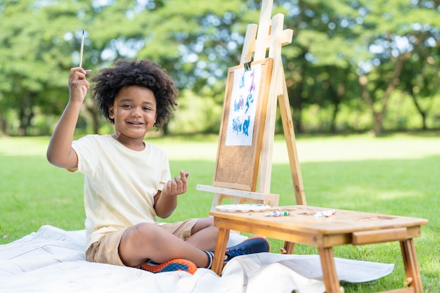 Garoto afro-americano mostra tinta de pincel com mini coração enquanto pinta aquarela em papel branco no parque