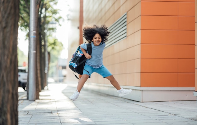 Garoto afro bonito pulando feliz com a mochila saindo da escola