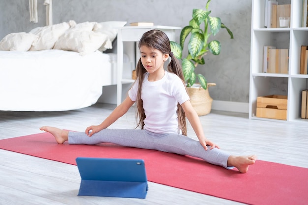 Garoto árabe fazendo alongamento on-line com tablet no tapete de fitness em casa no apartamento moderno