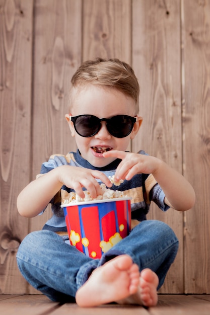 Garoto bonito bebê menino 2-3 anos de idade, óculos de cinema 3d imax segurando balde para pipoca, comendo fast-food em fundo de madeira. Conceito de estilo de vida de infância de crianças.