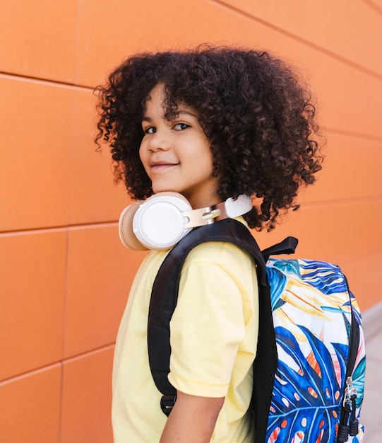 Foto garoto bonito com cabelo afro, indo para a pré-escola, com mochila e fones de ouvido, foto vertical
