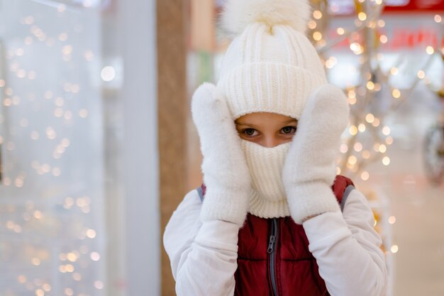 Garoto bonito com chapéu branco bobble e lenço no shopping perto da vitrine luzes brilhantes desfocadas no fundo