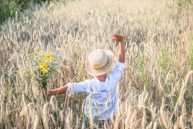 Garoto bonito com chapéu de palha e margaridas nas mãos, caminhando no campo de trigo de centeio