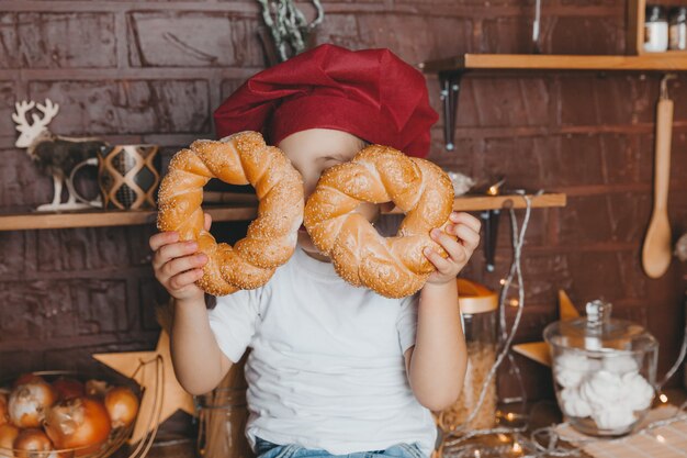 Garoto bonito com um chapéu de chef se senta na cozinha e segura pãezinhos perto de seu rosto.