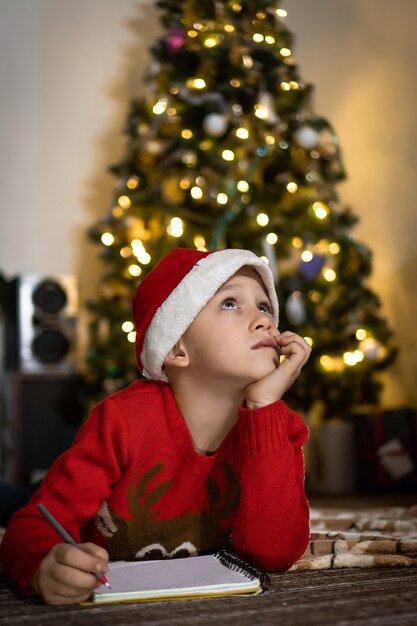 Garoto bonito de suéter vermelho e chapéu de papai noel vermelho escreve uma carta para o papai noel