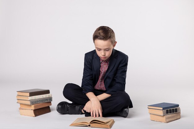 Garoto bonito de terno sentado e lendo um livro em uma pilha de livros de fundo branco