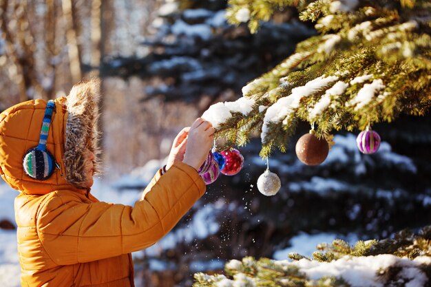 Foto garoto bonito em pano quente e chapéu pegando bola de natal em winter park. as crianças brincam ao ar livre no bosque nevado. crianças pegam bolas de natal.