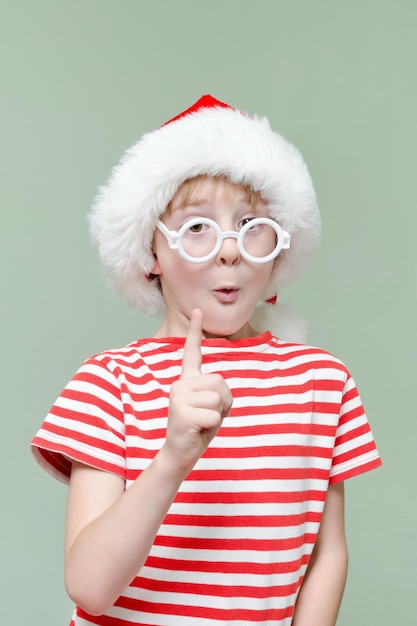 Garoto bonito em um chapéu de Papai Noel e óculos levantou o dedo