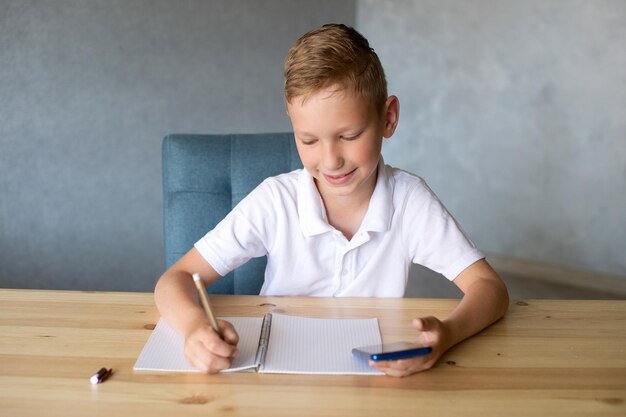 Garoto bonito escreve em um caderno aberto e olha para o telefone