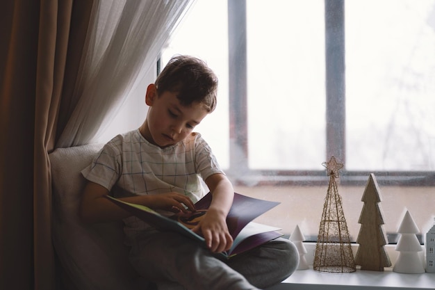 Garoto bonito está lendo um livro sentado no peitoril da janela perto da janela casa aconchegante com árvores de natal decorativas doce lar estilo de vida de férias de inverno