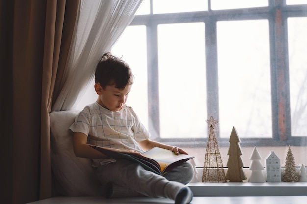 Garoto bonito está lendo um livro sentado no peitoril da janela perto da janela Casa aconchegante com árvores de Natal decorativas Doce lar Estilo de vida de férias de inverno