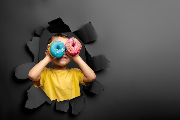 Garoto bonito feliz está se divertindo jogou com donuts na parede preta.