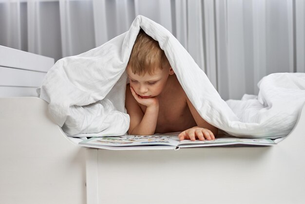 Garoto bonito lendo um livro em uma cama branca