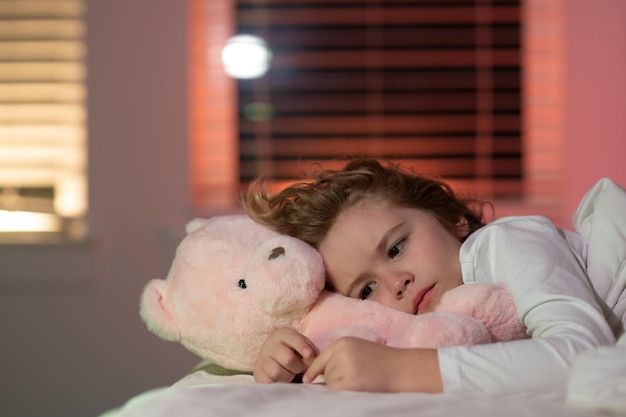 Garoto bonito tentando adormecer com ursinho de pelúcia vai dormir à noite no corte do quarto