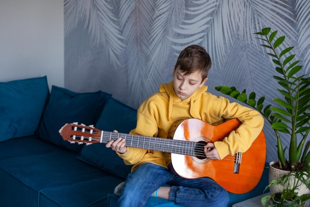 Garoto bonito tocando violão vestido jeans casual e moletom amarelo no sofá aconchegante em casa