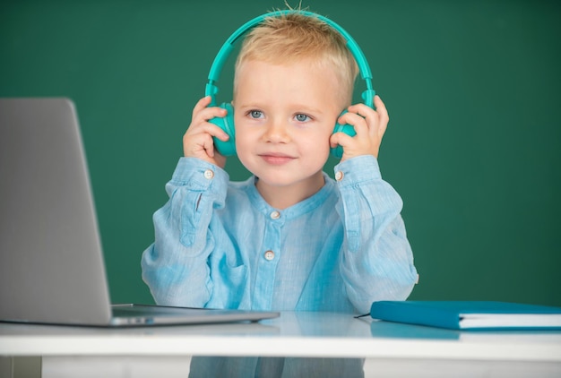 Garoto bonito usando fones de ouvido estuda com laptop em sala de aula ouvindo aula de áudio