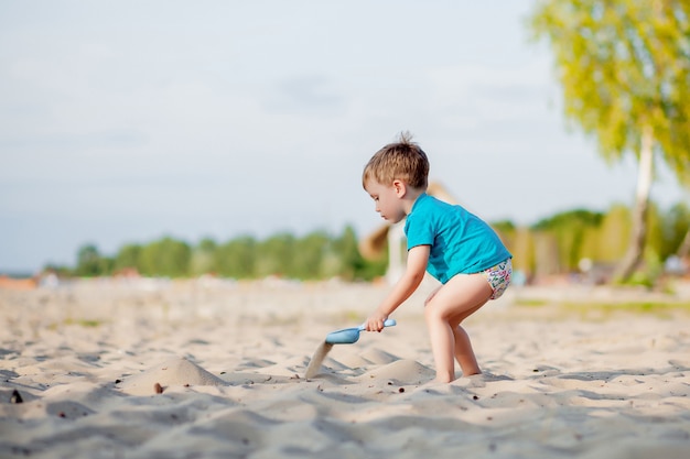 Garoto brincando na praia. Brincadeira no mar em férias de verão. Brinquedos de areia e água, proteção solar para crianças pequenas. Rapaz pequeno que escava a areia, construindo o castelo na costa do oceano.