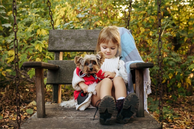 Garoto com cachorro engraçado está sentado em uma grande cadeira de madeira no jardim. Criança do sexo feminino com cachorrinho posa no quintal. Infância feliz