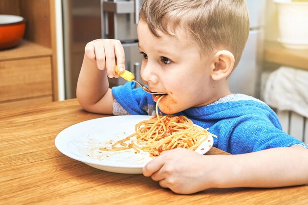 Garoto com olhos grandes come macarrão delicioso e desleixado com garfo de bebê amarelo sentado na cozinha moderna