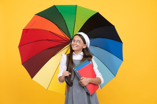 Garoto de boina com guarda-chuva de arco-íris temporada de outono previsão de tempo chuvoso de volta à escola proteção contra chuva vívida garota feliz de óculos segura notebook educação adolescente sob guarda-sol colorido