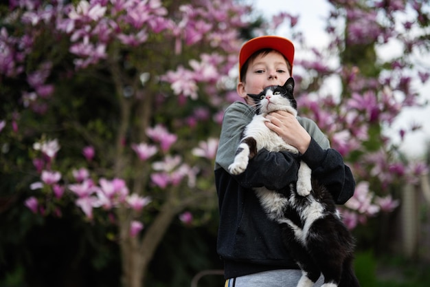 Garoto de boné segura gato nas mãos contra um belo dia de primavera perto da árvore florescente de magnólia