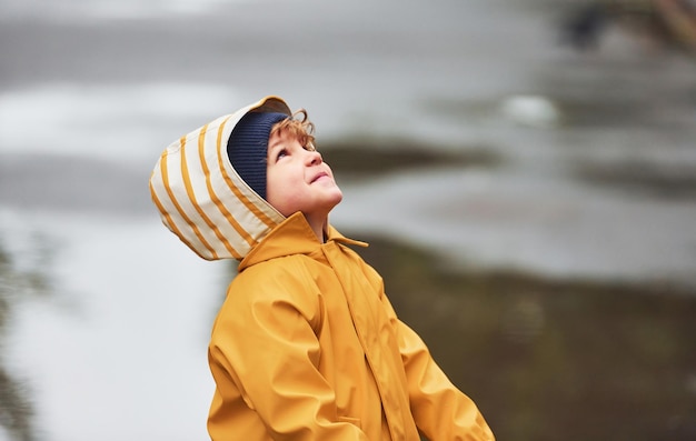 Garoto de capa impermeável amarela e botas brincando ao ar livre depois da chuva