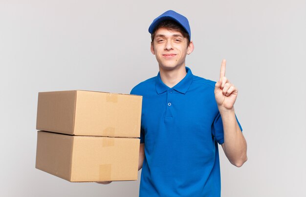 Garoto de entrega de pacote jovem sorrindo e parecendo amigável, mostrando o número um ou o primeiro com a mão para a frente, em contagem regressiva