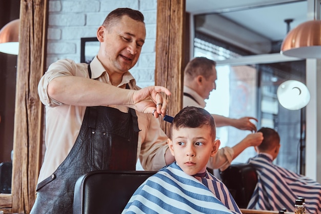 Garoto de escola atraente está cortando o cabelo na moda do cabeleireiro maduro no salão de cabeleireiro da moda.