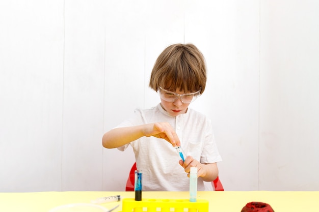 Garoto de óculos de segurança e camiseta branca fazendo experiências no laboratório químico. Garotinho com frascos para química. Educação escolar em casa.