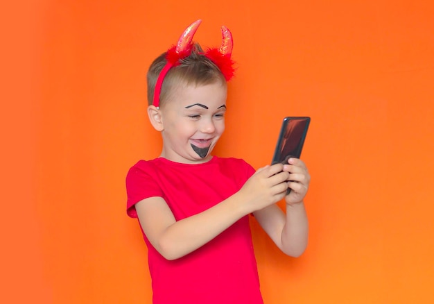 Garoto engraçado em uma fantasia de halloween tira uma selfie em um celular em um fundo laranja o garoto liga para o telefone para dizer que está pronto para o feriado de doces