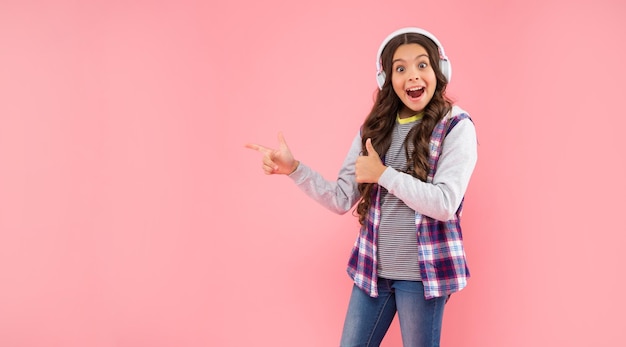 Garoto espantado ouvindo música em fones de ouvido aponta o dedo no fundo rosa com tecnologia de espaço de cópia