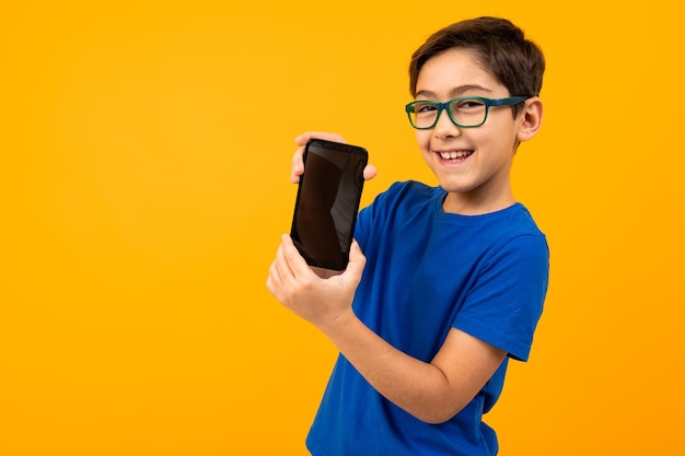 Garoto esperto de óculos e uma camiseta azul segura o telefone com a tela para a frente com um layout em um fundo amarelo com espaço de cópia.