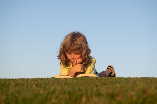 Garoto esperto lendo livro no parque ao ar livre um jovem garoto loiro está deitado na grama verde ao ar livre r