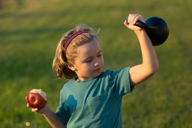 Garoto esportivo com maçã e kettlebell ao ar livre no parque de verão Atividades esportivas de lazer com crianças Menino loiro segurando kettlebell Exercícios esportivos para crianças Criança engraçada levantando os halteres
