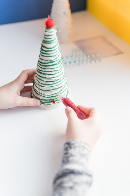 Garoto está mostrando uma decoração da árvore de Natal. Fabricação de árvore de Natal artesanal criativa. Conceito de DIY infantil. Fazendo decoração de brinquedos de natal. Decore o brinquedo com strass. Árvore de Natal ecológica