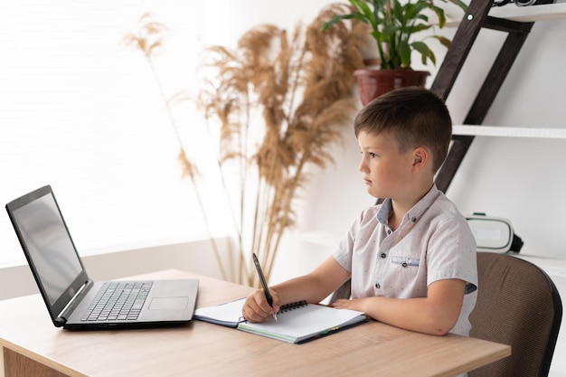 Garoto estudante estudando em casa fazendo lição de casa com um laptop anotando a tarefa em um caderno ou bloco de notas Estudante do ensino fundamental júnior estuda em casa Conceito de educação e aprendizagem à distância