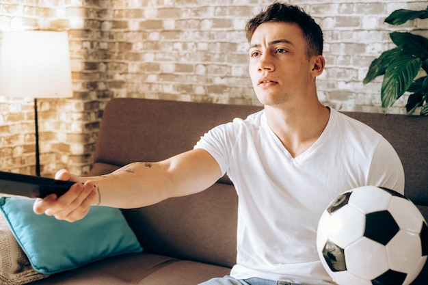 Garoto fã assistindo futebol na TV segurando uma bola de futebol decepcionado pelo gol perdido de seu time