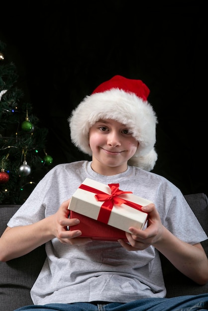 Garoto feliz com chapéu de Papai Noel abre caixa de presentes sentado no sofá contra o fundo da árvore de Natal Presentes de Ano Novo Quadro vertical