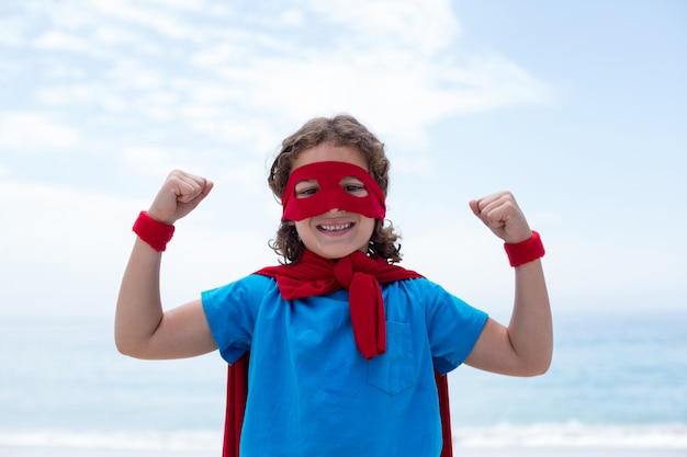 Foto garoto feliz em traje de super-heróis, flexionando os músculos na beira-mar