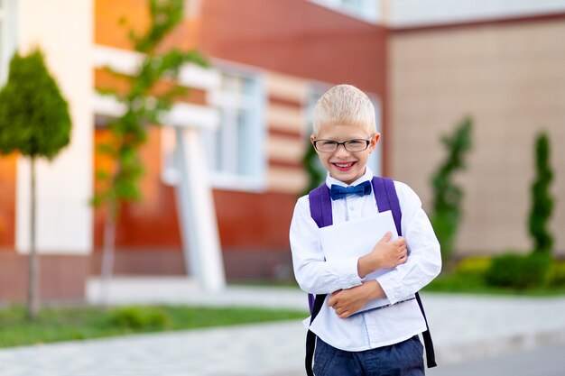 Garoto feliz garoto loiro de óculos com uma mochila e um livro branco fica na escola e ri. Dia de conhecimento