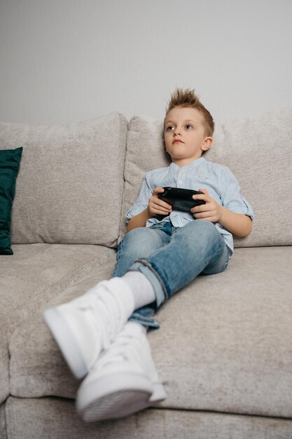 Garoto feliz jogando videogame segurando o controlador de jogo sentado no treinador na sala de estar