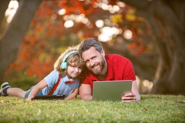 Garoto feliz ouve música em fones de ouvido. vídeo-aula de webinar. educação on-line no laptop. tutoria. pai e filho usam tecnologia de comunicação moderna no parque. blog da família. infância e paternidade.