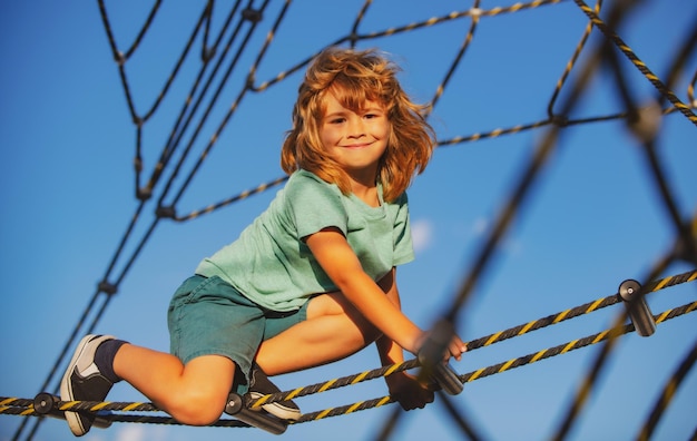 Garoto fofo e sorridente subindo na rede no parque infantil parque de cordas cara de crianças engraçadas
