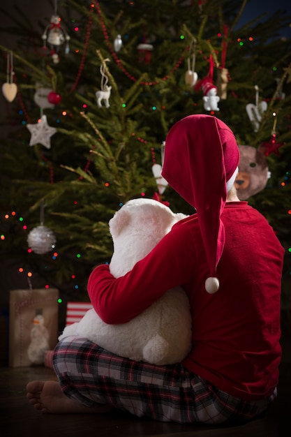 Garoto garoto de pijama sentado com seu brinquedo de urso perto de uma árvore de Natal e curtindo a celebração