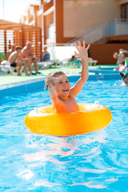 Garoto garoto jogando piscina ao ar livre do resort. em um círculo amarelo inflável com uma bola. As crianças brincam com brinquedos aquáticos. Espirrando ao redor. Esfrie o calor.