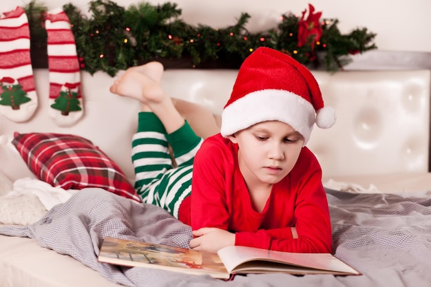 Garoto garoto no chapéu de Papai Noel e pijama de Natal lendo um livro.